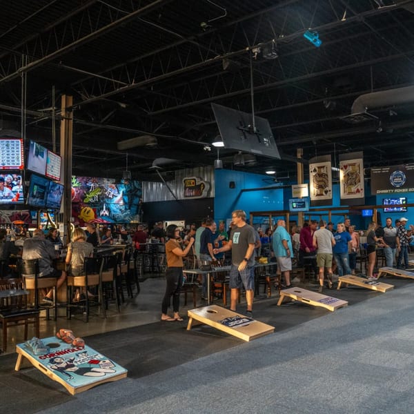 Patrons socialize and play cornhole at One Eyed Jacks bar-and-grill, with arcade-themed decor and TVs.
