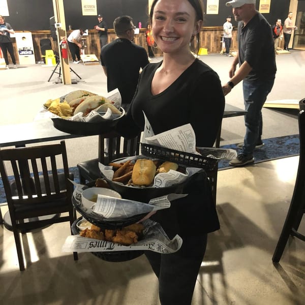 Smiling server carrying trays of One Eyed Jacks bar food, with people playing cornhole in the background.