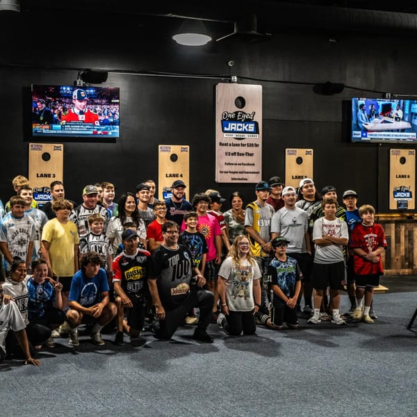 A large group of adults and children pose for a photo in front of cornhole boards at One Eyed Jacks.
