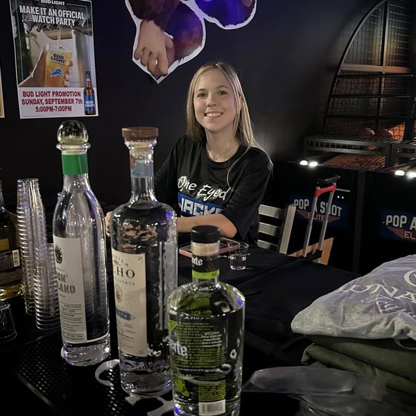 A smiling woman in a One Eyed Jacks shirt sits behind a bar with liquor bottles and plastic cups.