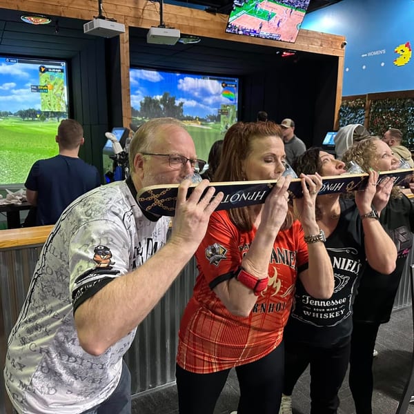 Four adults take shots from a ski, with golf simulators and a Pac-Man sign in the background.