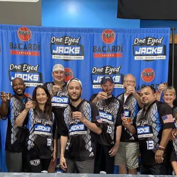 A group of people in One Eyed Jacks shirts toast drinks in front of a branded banner.