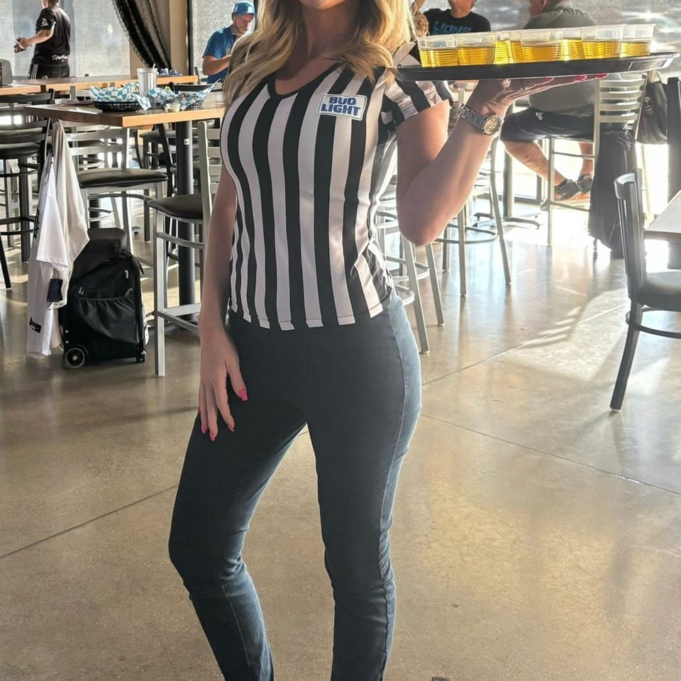 A server in a striped shirt with a "Bud Light" patch carries a tray of beer shots at One Eyed Jacks.