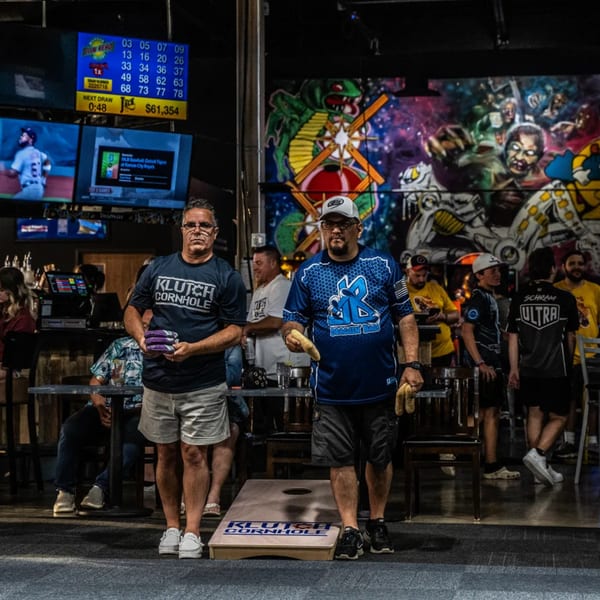 Two men playing cornhole at One Eyed Jacks, with a vibrant mural and TVs in the background.