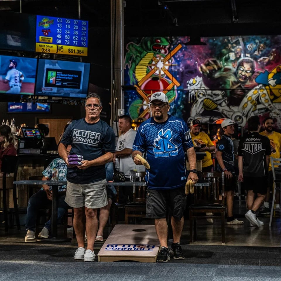 Two men playing cornhole at One Eyed Jacks, with a vibrant mural and TVs in the background.
