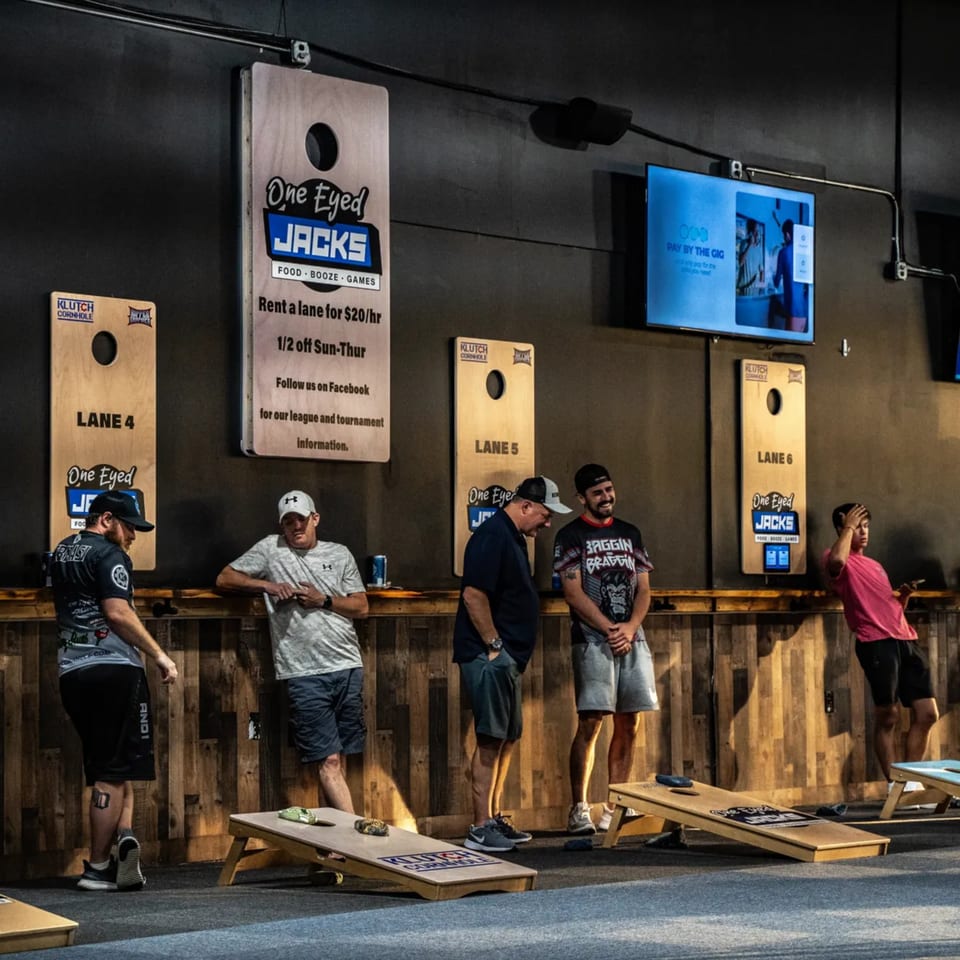 Men play cornhole at One Eyed Jacks, a bar-and-grill with games and screens.