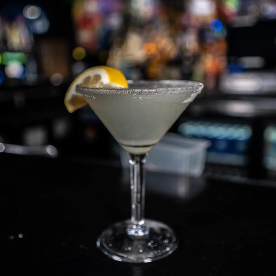 A margarita with a salted rim and lemon wedge on a dark bar with blurred lights in the background.