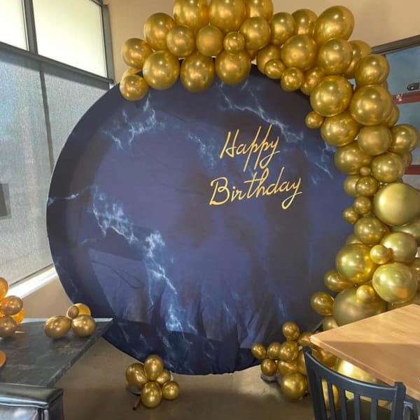 A circular blue marble-patterned backdrop with "Happy Birthday" in gold, framed by gold balloons.