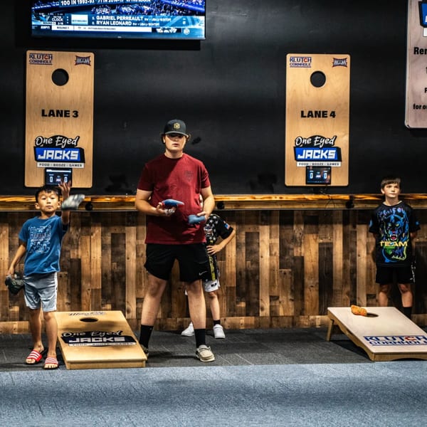 Kids playing cornhole at One Eyed Jacks, a bar-and-grill with games and a TV screen.
