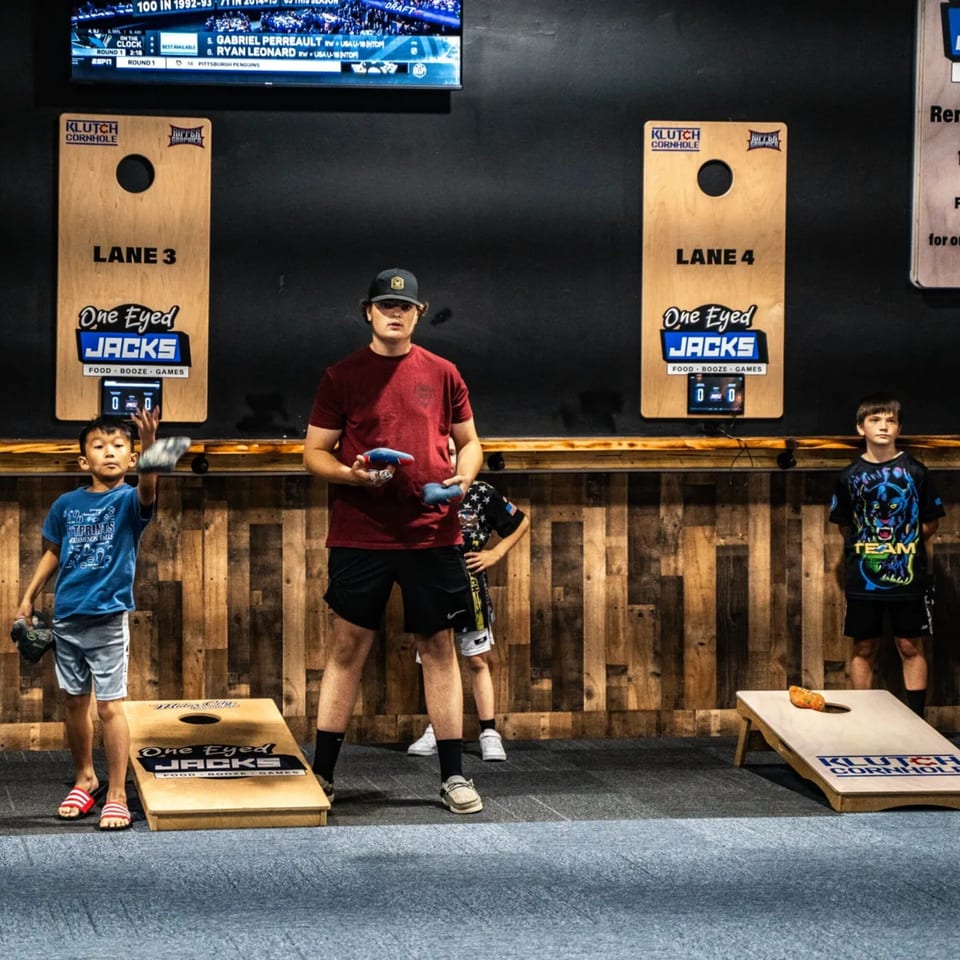 Kids playing cornhole at One Eyed Jacks, a bar-and-grill with games and a TV screen.