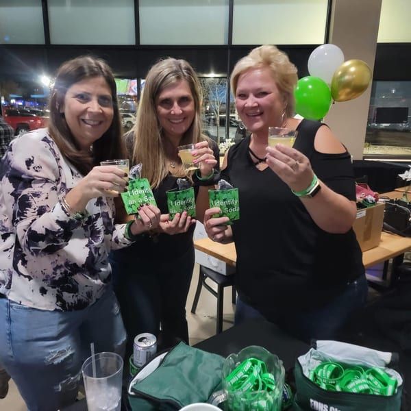 Three women smiling and holding drinks, with green, white, and gold balloons in the background.