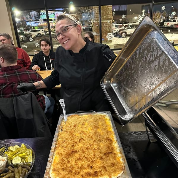 A smiling chef in black uniform serves a large pan of mac and cheese at One Eyed Jacks.