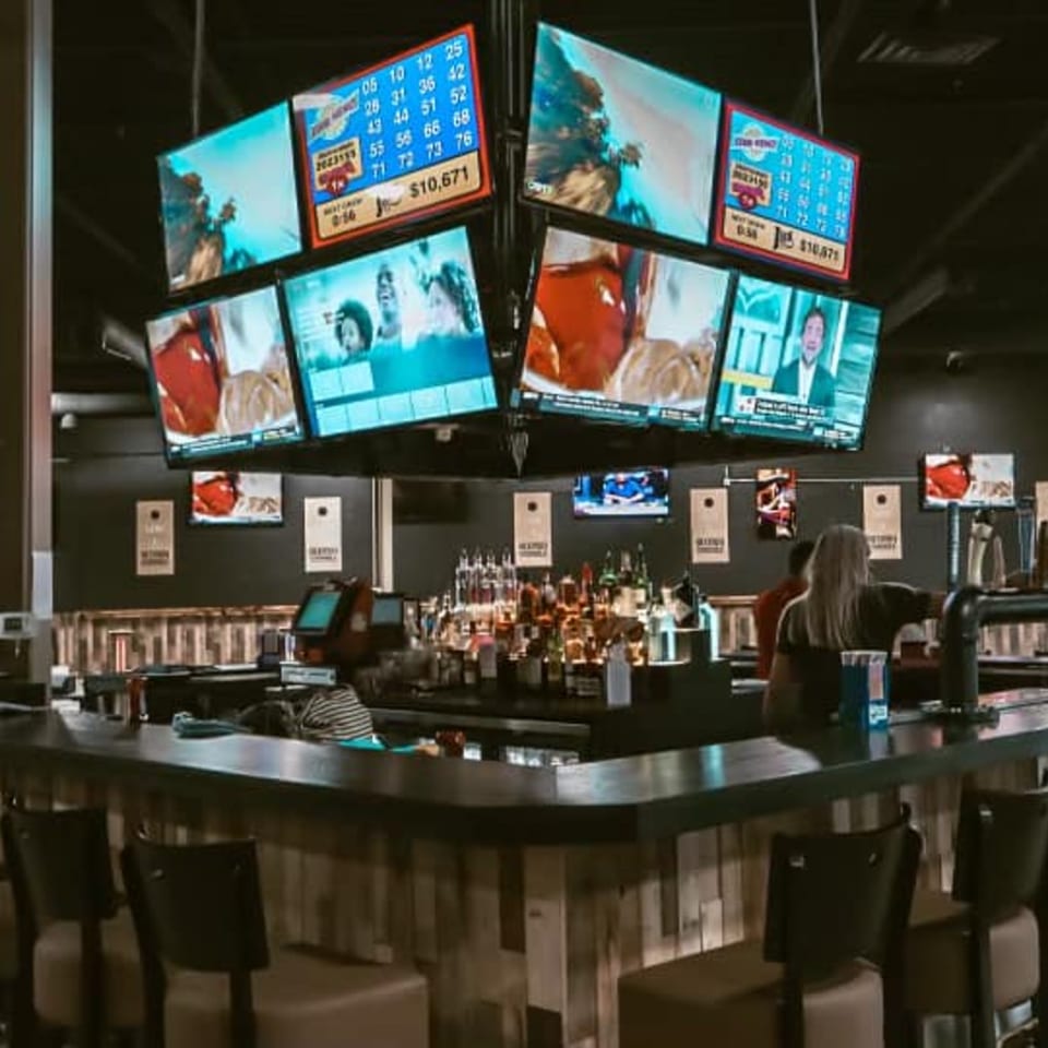 A bar at One Eyed Jacks with multiple TVs showing sports and a lottery, with patrons seated.