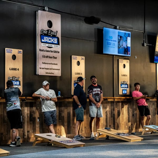 Men playing cornhole at One Eyed Jacks bar-and-grill, with cornhole boards and screens visible.