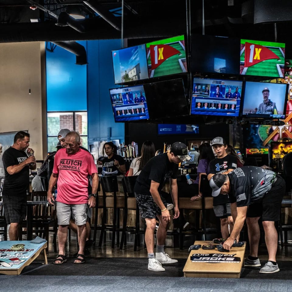 Patrons playing cornhole and watching sports on multiple TVs at One Eyed Jacks bar-and-grill.