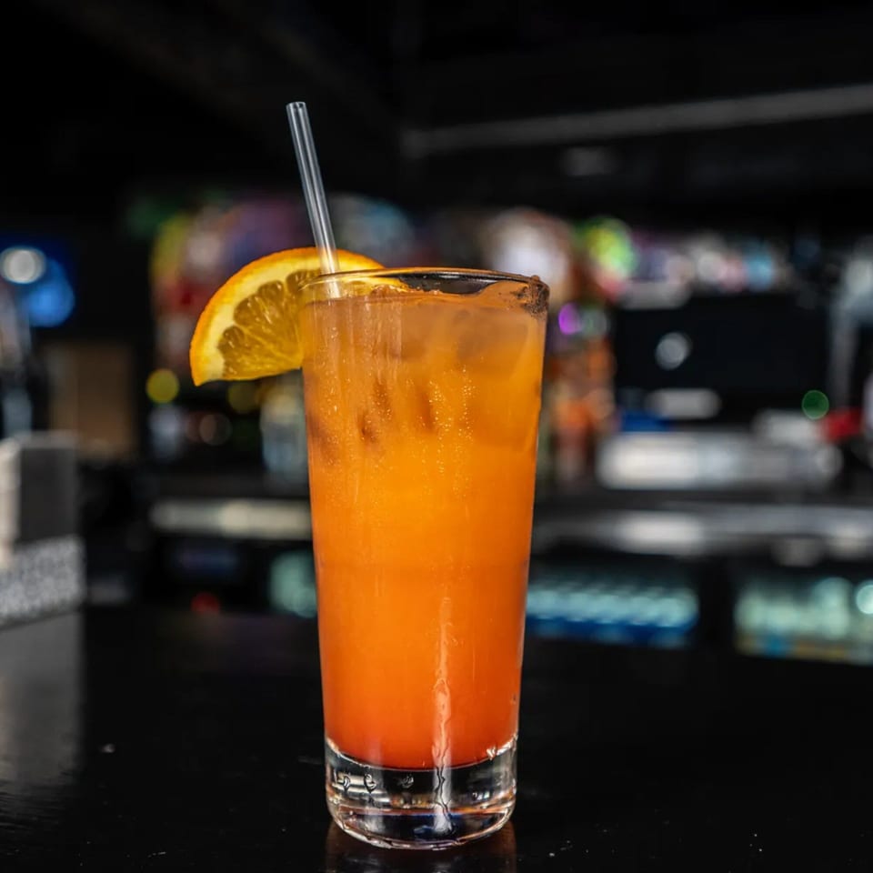 A vibrant orange cocktail with an orange slice and straw sits on a dark bar at One Eyed Jacks.