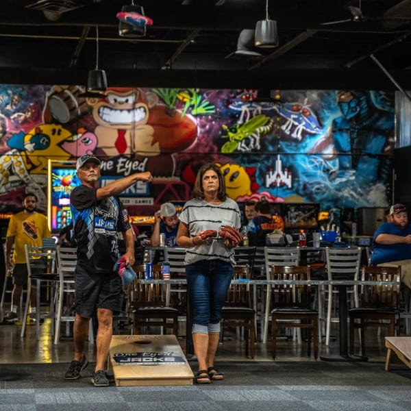 People playing cornhole inside One Eyed Jacks bar-&-grill, with colorful arcade-themed murals in the background.