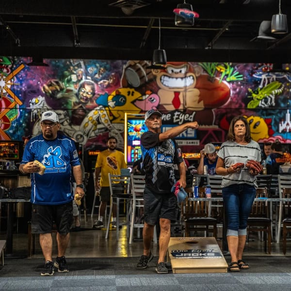 People playing cornhole at One Eyed Jacks bar-&-grill, with a vibrant mural in the background.