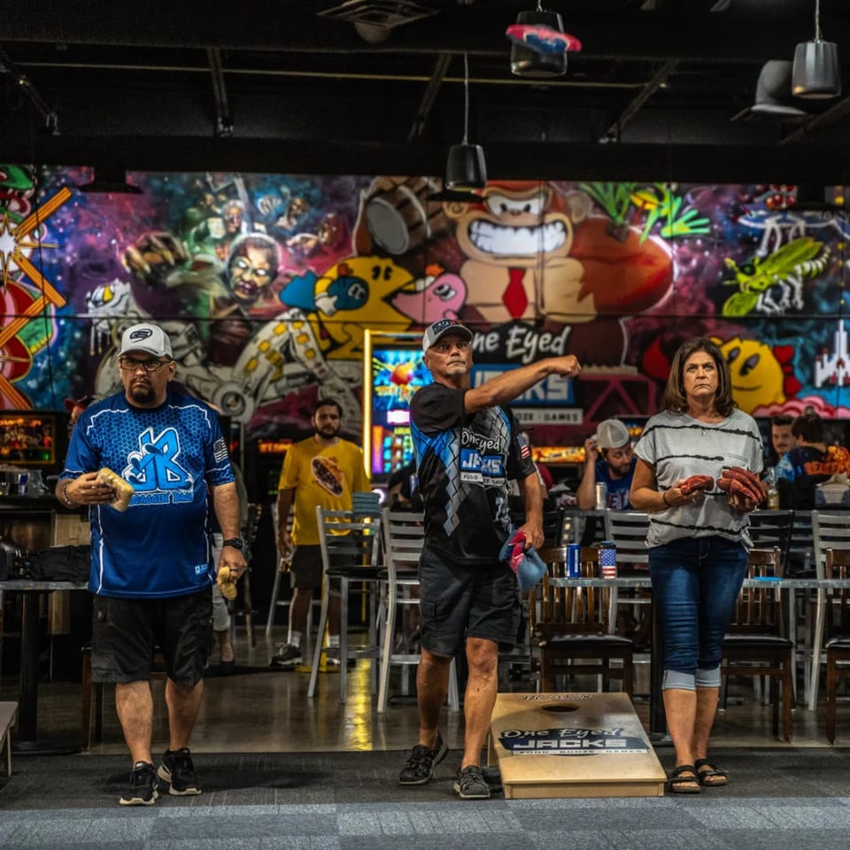 People playing cornhole at One Eyed Jacks bar-&-grill, with a vibrant mural in the background.