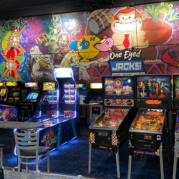 A vibrant mural of classic video game characters adorns the wall above arcade games and pinball machines at One Eyed Jacks.