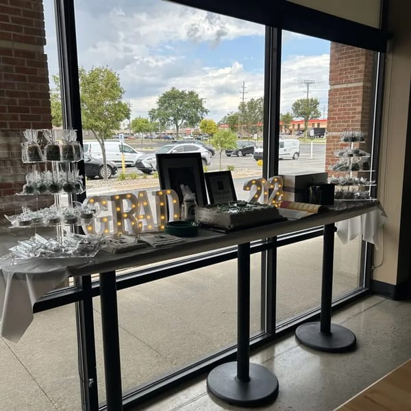 A graduation party setup with a cake, "GRAD 22" lights, and wrapped treats on a table by a window.