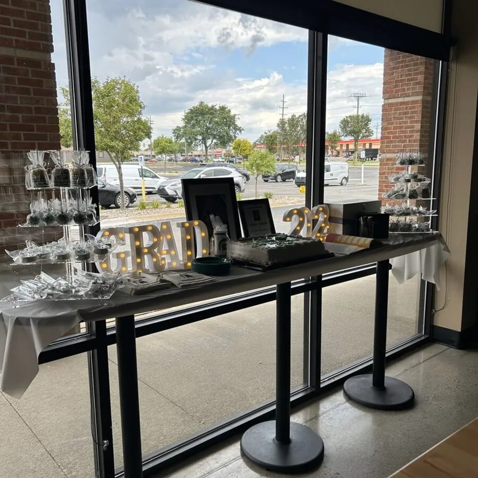 A graduation party setup with a cake, "GRAD 22" lights, and wrapped treats on a table by a window.