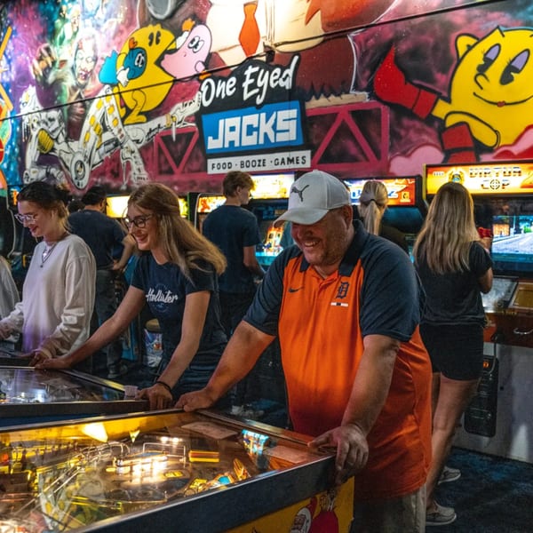 People playing pinball and arcade games at One Eyed Jacks, with colorful graffiti on the wall.