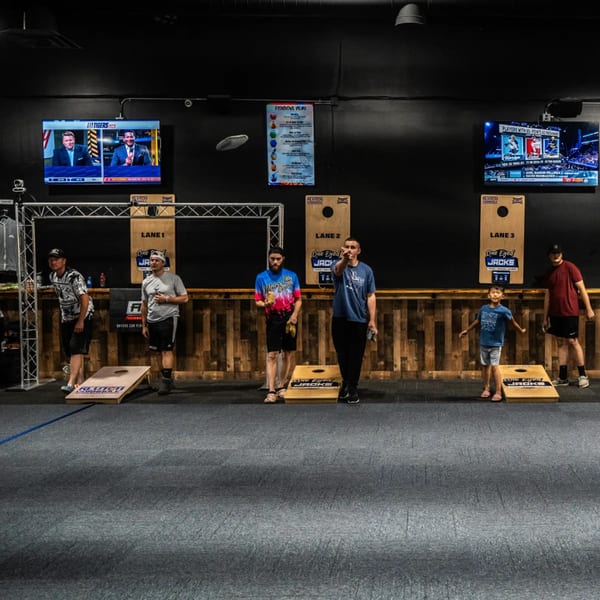 People playing cornhole at One Eyed Jacks, with TVs showing sports and a menu on the wall.