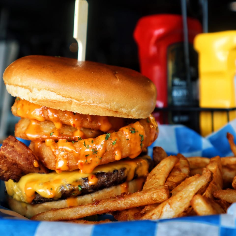 A One Eyed Jacks burger with onion rings, cheese, and sauce, served with fries in a blue-and-white basket.
