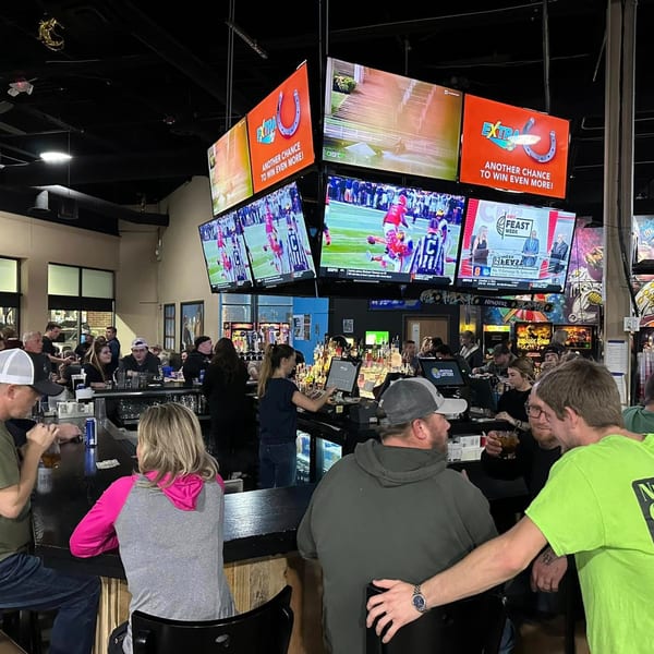 Patrons socialize at the bar of One Eyed Jacks, with multiple TVs displaying sports and advertisements.