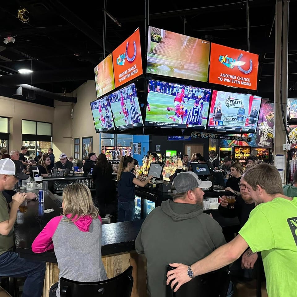 Patrons socialize at the bar of One Eyed Jacks, with multiple TVs displaying sports and advertisements.