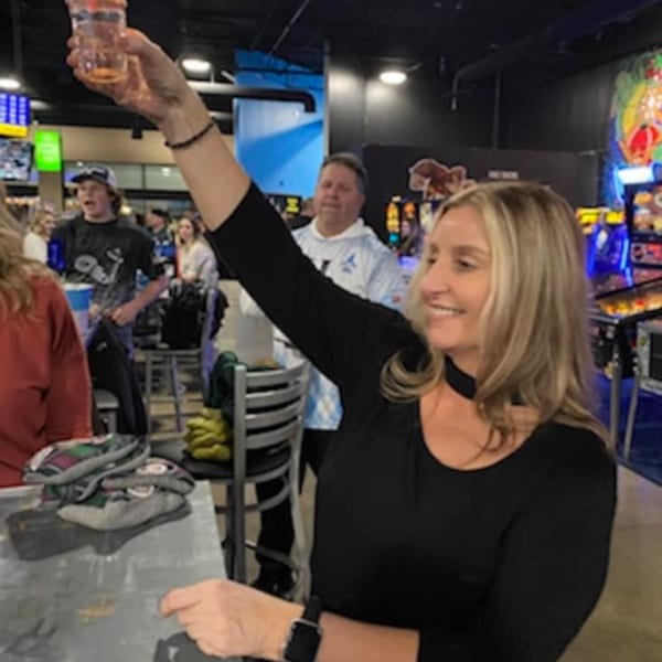 A woman in a bar-and-grill raises a drink in a toast, smiling amidst other patrons.