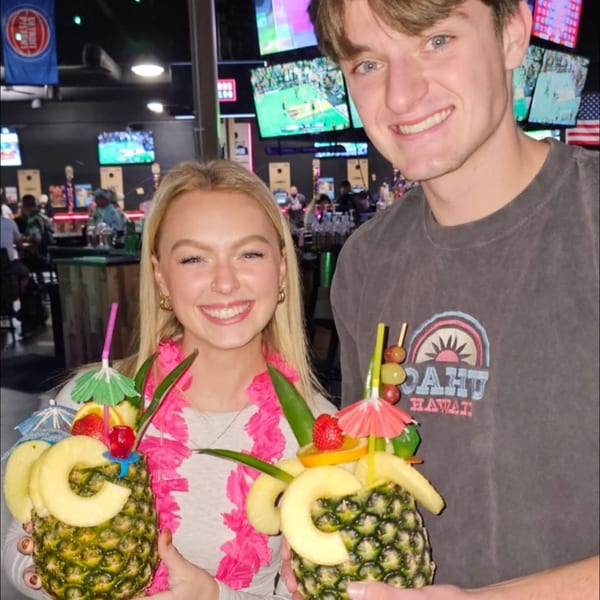 Two smiling people holding pineapple drinks with fruit garnishes and umbrellas at One Eyed Jacks.