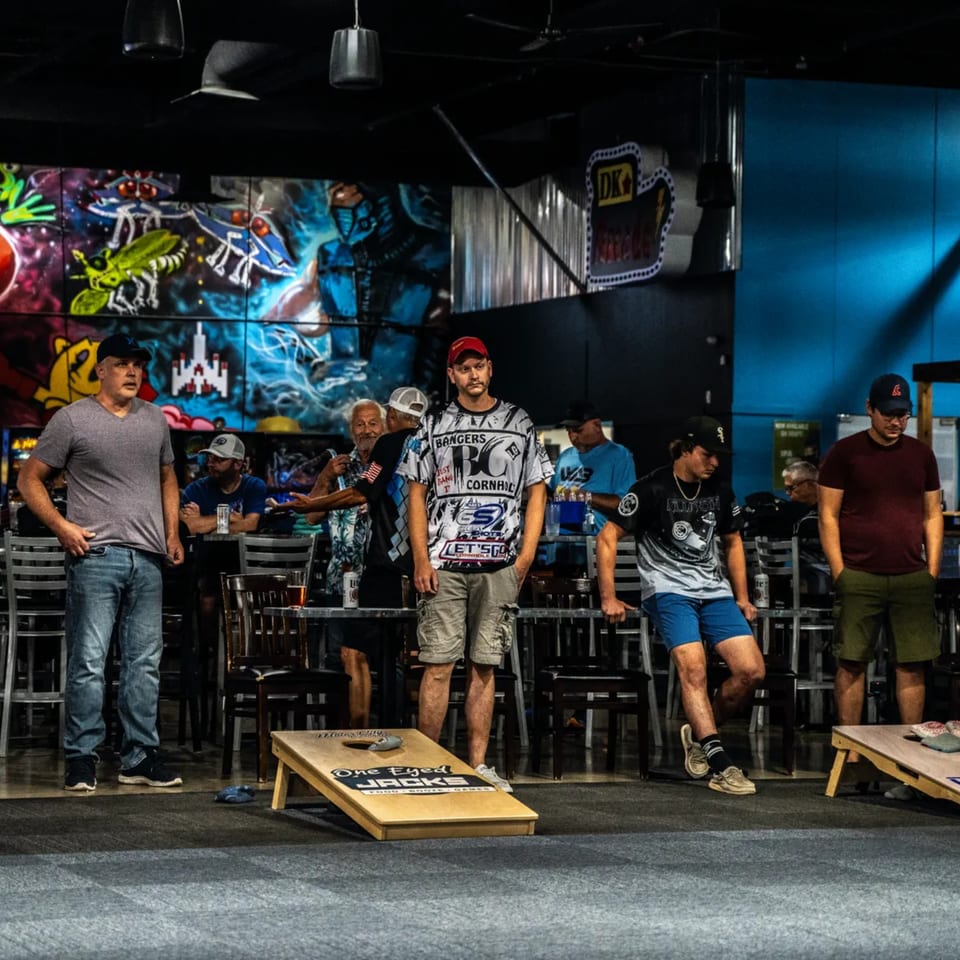 Men playing cornhole inside One Eyed Jacks bar-and-grill with colorful graffiti walls.