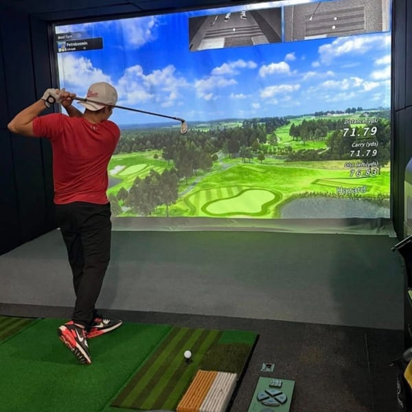 A person in a red shirt and white hat swings a golf club at a virtual golf simulator.