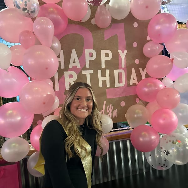 Smiling woman in a black top and gold sash in front of a "Happy Birthday" sign and pink balloons.
