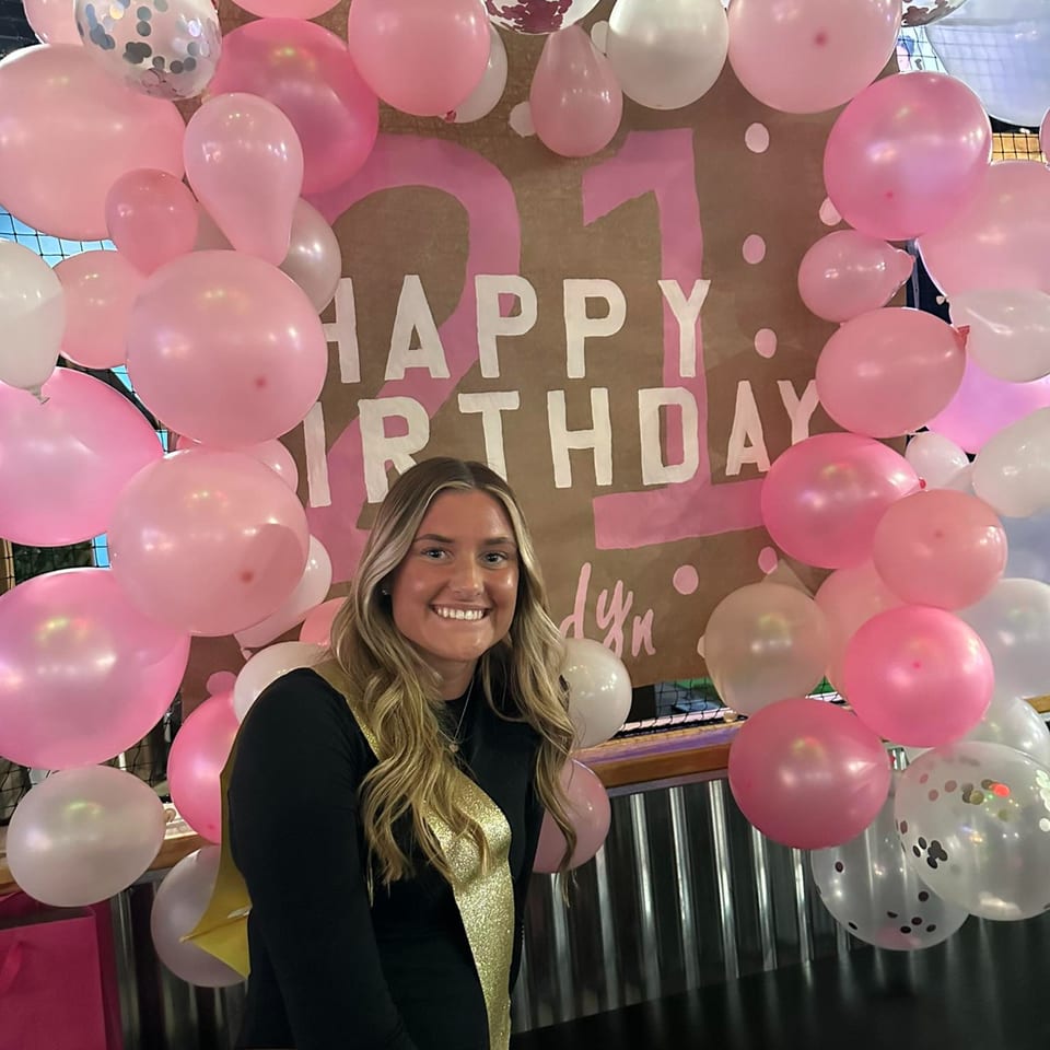 Smiling woman in a black top and gold sash in front of a "Happy Birthday" sign and pink balloons.