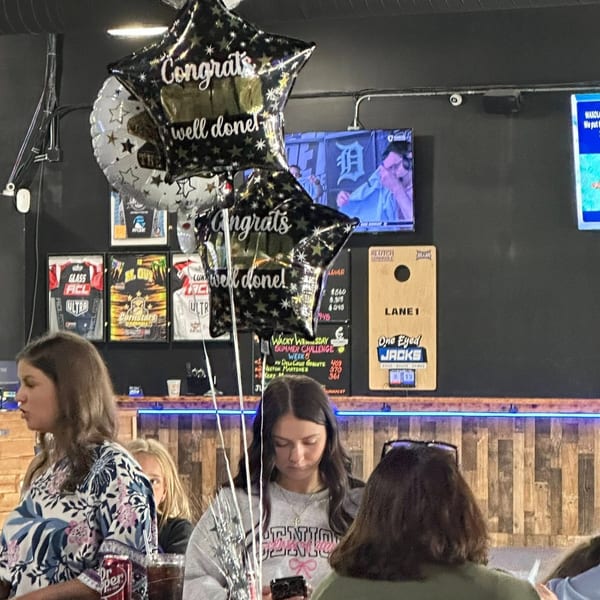 Patrons at One Eyed Jacks bar-&-grill, with "Congrats well done!" balloons and sports screens.