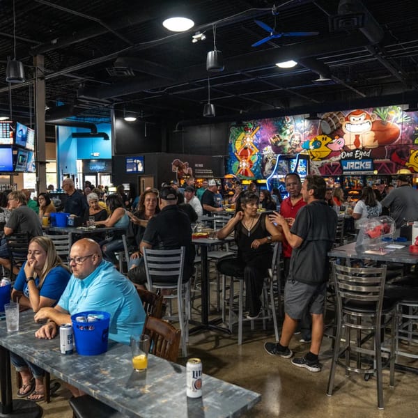 Patrons socialize at high-top tables in a lively bar-and-grill with arcade-themed murals and TVs.